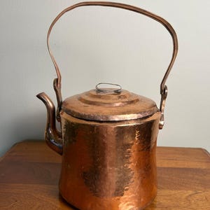 May include: A hammered copper kettle with a cylindrical body, a curved spout, and a tall, arched handle. The kettle features a circular lid with a small metal loop handle on top. The surface has a reflective, metallic finish with visible texture.