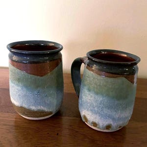 May include: Two ceramic mugs featuring a multi-colored glaze with horizontal bands of dark blue, brown, sage green, and light blue. The mugs have a cylindrical shape with a slightly flared rim and one mug includes a curved handle on the side.