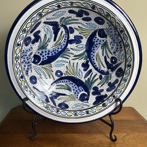 May include: A decorative ceramic plate featuring a hand-painted design of three blue fish swimming among aquatic plants. The plate has a white background with a blue border and is displayed on a black metal stand.