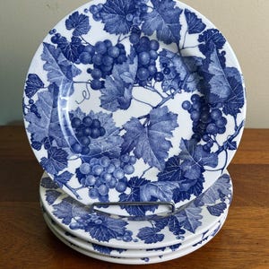 May include: A set of four white ceramic plates with a blue grape and leaf design. The plates are stacked on a metal rack, showcasing the detailed botanical pattern. The plates are suitable for serving food.
