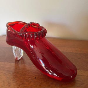 May include: A decorative glass shoe figurine in a vibrant translucent red color. The shoe features a rounded toe, a textured strap across the top opening, and a clear, faceted glass heel that provides support for the structure of the object.