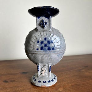 May include: A gray ceramic vase with a circular body and a dark blue glazed rim. The surface features embossed geometric patterns, including a central grid of blue squares, radiating lines, and a zigzag motif on the base, all in a matte finish.