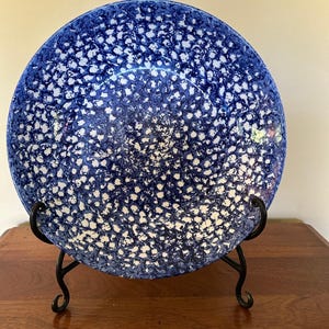May include: A round ceramic plate featuring a speckled blue and white pattern. The plate is displayed upright on a black metal stand, showcasing a dense, mottled design across its entire surface with a smooth, glossy finish and a circular rim.