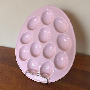 May include: A light pink ceramic egg serving platter shaped like a large egg, featuring twelve oval-shaped indentations arranged in a grid pattern. The platter is held upright by a thin, silver-toned metal wire stand that supports the base.