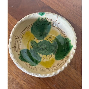 May include: A handmade ceramic bowl with a cream-colored base and a decorative interior. The bowl features green and yellow painted accents, with a central green floral design. The rim has a textured, wavy edge and the bowl is sitting on a wooden surface.