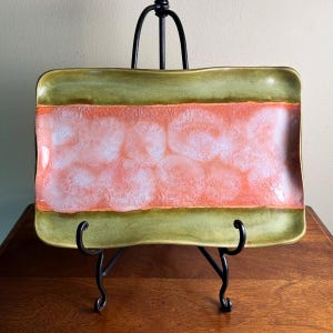 May include: A rectangular ceramic tray featuring a wide central band of mottled coral and white glaze, bordered by solid olive green edges. The tray has slightly curved, raised sides and is displayed on a black metal wire stand.
