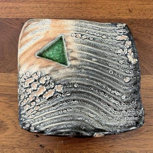 May include: A square ceramic plate with a textured surface featuring a gradient from light beige to dark charcoal gray. The surface has a wavy, crackled pattern and a single, prominent, smooth, bright green triangular inlay in the upper left.