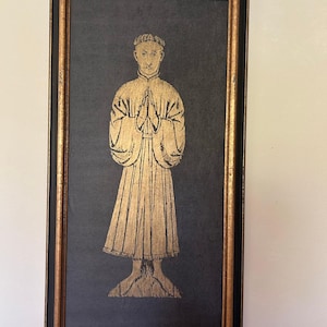 May include: A framed artwork featuring a gold-colored illustration of a figure in a robe with hands clasped in prayer. The artwork is set against a dark background and framed in black with gold trim.