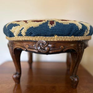 May include: A round footstool featuring a dark blue and cream needlepoint floral pattern on the top cushion. The base is constructed from dark brown carved wood with curved legs and a gold-colored braided trim bordering the upholstered seat.