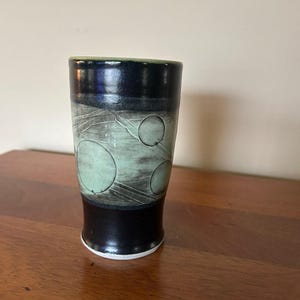 May include: A handmade ceramic tumbler with a black base and rim, featuring a light green and gray central band with circular designs. The top rim is dark blue and the interior is green. The tumbler sits on a wooden surface.