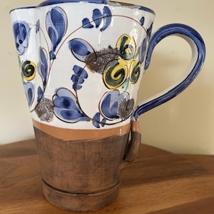 May include: A ceramic pitcher with a white background, adorned with blue floral patterns and yellow accents. The handle and rim are blue, and the lower portion is brown. The pitcher is sitting on a wooden surface.
