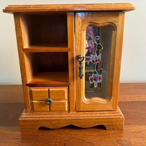 May include: A wooden jewelry armoire with a light brown finish, featuring two open shelves, a small drawer with a round knob, and a hinged door with a glass panel. The glass panel displays a painted floral design and a metal handle.