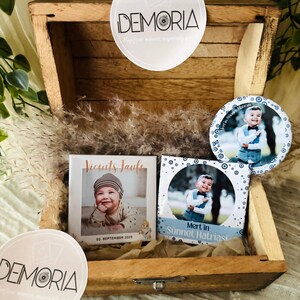 May include: A wooden box holds personalized photo magnets and round stickers. The magnets feature baby photos with text. The stickers display the business name "DEMORIA". The box is filled with decorative dried grass.