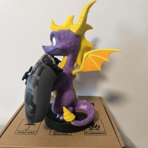 May include: A purple dragon figurine with yellow horns, wings, and accents, holding a camouflage-patterned game controller. The figurine is on a black base, and the box below has text that reads "KEEP DRY" and "FLAMMABLE".