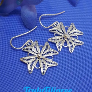 Può includere: Un paio di orecchini in filigrana d'argento a forma di fiore con un design delicato e intricato. Gli orecchini sono su uno sfondo blu.