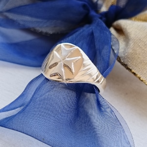 May include: A silver ring with a Maltese cross design. The ring is on a blue and gold fabric background.