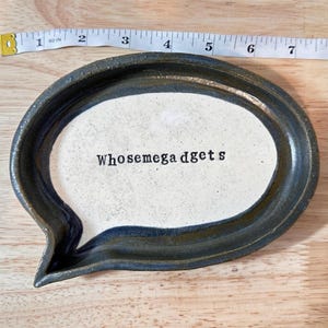 Könnte beinhalten: Eine sprechblasenförmige Keramikschale mit dunkelgrauem Rand und gesprenkeltem, cremeweißem Zentrum. Der Text "Whosemegadgets" ist in der Mitte schwarz aufgedruckt. Ein gelbes Maßband zeigt eine Breite von etwa 20 cm an.