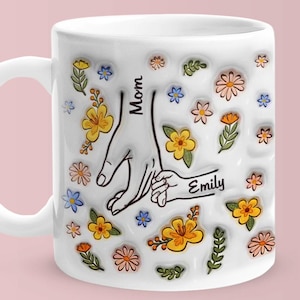 Puede incluir: Taza de cerámica blanca con diseño floral y las palabras "Mom" y "Emily". La taza presenta una ilustración de manos entrelazadas, rodeada de flores coloridas en amarillo, naranja y azul. Un regalo para el Día de la Madre.
