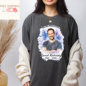 May include: Dark gray t-shirt featuring a portrait of a man with the text "Remembering Leonid Radvinsky 1982-2026". The portrait is surrounded by a watercolor-style design in shades of blue and purple. The shirt is a casual style.