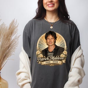 May include: Dark gray t-shirt featuring a portrait of Patrick Muldoon within a circular design with a vintage aesthetic. The design includes the text "Patrick Muldoon" and the years "1968 - 2026". The shirt has a relaxed fit.