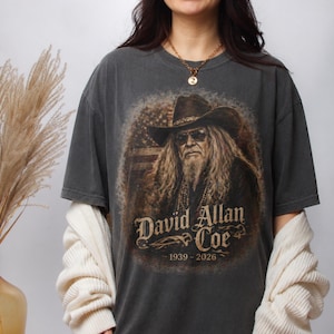 May include: Dark gray t-shirt featuring a portrait of David Allan Coe wearing a cowboy hat and sunglasses, with the American flag in the background. The text "David Allan Coe" and the years "1939-2026" are printed below the portrait.