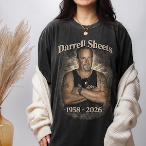 May include: Dark gray t-shirt featuring a portrait of Darrell Sheets with the text "Darrell Sheets" above the image and the years "1958-2026" below. The shirt has a vintage, worn look.