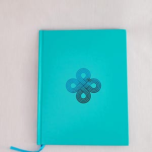 May include: A teal-colored journal with a geometric design on the cover. The design is a series of interlocking loops in shades of blue and black. A matching teal ribbon bookmark extends from the top edge.