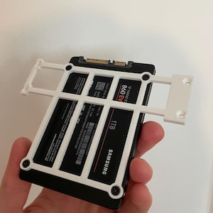 May include: A black Samsung 1TB SSD with white mounting bracket. The SSD has the text "860 EVO" and "1TB" printed on it. The white bracket is secured with black screws. The SSD is held in a person's hand.