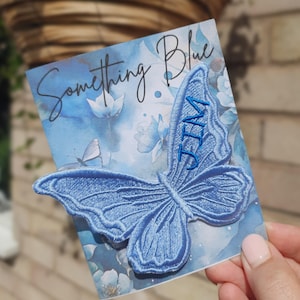 Puede incluir: Un parche bordado de mariposa en color azul claro con el nombre JIM cosido en hilo azul oscuro. El parche está sujeto a una tarjeta rectangular con un diseño floral de acuarela en tonos azules y el texto Something Blue en la parte superior.