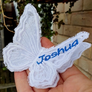 Puede incluir: Una mariposa de encaje blanco con el nombre Joshua bordado en hilo azul en el ala inferior. La mariposa presenta intrincados detalles de encaje calado en todas sus alas, creando una apariencia delicada y texturizada.