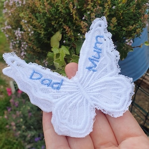 Op de afbeelding: Een witte kanten vlinderapplicatie met lichtblauw geborduurde tekst op de vleugels. De linkervleugel toont het woord Dad en de rechtervleugel toont het woord Mum. Het kant heeft een verfijnde, geschulpte rand en een gedetailleerd stikpatroon.