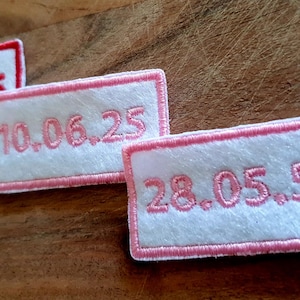 May include: Three rectangular embroidered patches with dates. The patches have a white background with pink or red borders. The dates are embroidered in pink or red thread. The patches are on a wooden surface.