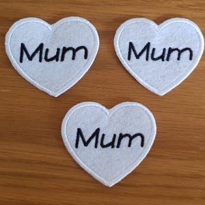 Può includere: Tre toppe bianche a forma di cuore con la scritta "Mum" ricamata in nero. Le toppe hanno un bordo bianco e sono disposte su una superficie di legno. Adatte per lavori di artigianato o come regalo.