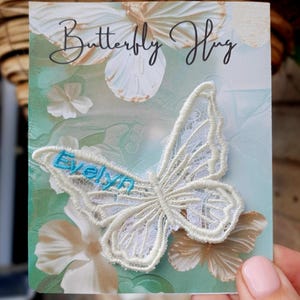Può includere: Una toppa a forma di farfalla in pizzo bianco con il nome Evelyn ricamato in filo azzurro, fissata su un cartoncino rettangolare con una grafica di farfalla e la scritta Butterfly Hug in carattere corsivo nero in alto.