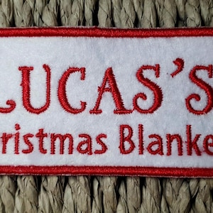 May include: A rectangular white patch with a red border and red embroidered text. The text reads "LUCAS'S Christmas Blanket". The patch is set against a woven, textured background.