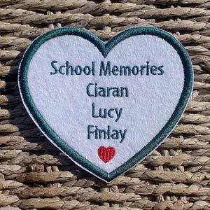 May include: A heart-shaped embroidered patch with the words "School Memories" and the names Ciaran, Lucy, and Finlay in teal. A small red heart is at the bottom. The patch has a white centre and a teal border.