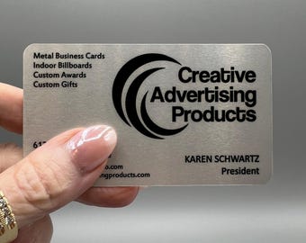 May include: A silver metal business card with black text and a logo for Creative Advertising Products. The card lists services such as metal business cards, indoor billboards, custom awards, and custom gifts. The card also includes the name Karen Schwartz, President.