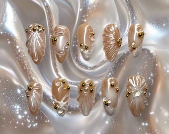 May include: A set of ten artificial nails with a pearlescent finish. Each nail features a unique design with gold-colored embellishments, including small beads and decorative accents. The nails have a light beige base color.