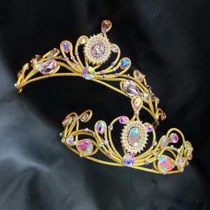 May include: Two gold-toned metal tiaras featuring intricate wirework designs. Each crown is adorned with iridescent, light-reflecting crystals and central oval-shaped stones surrounded by small white beads, creating a shimmering, ornate appearance.