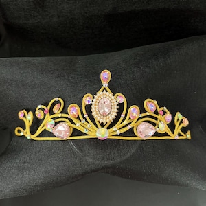 Puede incluir: Una tiara dorada con piedras preciosas rosas e iridiscentes. La tiara presenta una piedra preciosa rosa de forma ovalada en el centro, rodeada de piedras claras más pequeñas. El diseño incluye gemas rosas en forma de lágrima y gemas iridiscentes más pequeñas.