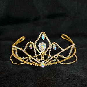 Puede incluir: Una tiara dorada adornada con piedras preciosas iridiscentes y transparentes. La corona presenta una gema central en forma de lágrima y gemas más pequeñas a lo largo de la banda. La tiara está sobre un fondo oscuro.
