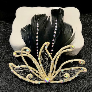 May include: A decorative hair accessory featuring a gold-toned filigree design with iridescent rhinestones and black feathers. The accessory is set against a white background and a black surface.