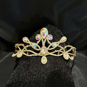 Puede incluir: Una tiara dorada con piedras preciosas iridiscentes y cristales transparentes. La tiara presenta un cristal central en forma de lágrima y está colocada sobre un fondo negro. El diseño incluye líneas curvas y detalles florales.
