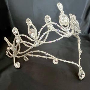 Puede incluir: Una tiara plateada con cristales transparentes y brillantes. El diseño presenta líneas curvas y elementos decorativos, con cristales más grandes en las puntas y extremos. Ideal para ocasiones formales.