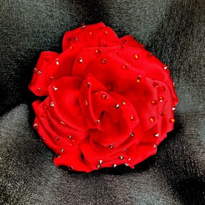 Op de afbeelding: Een felrode stoffen roos, versierd met sprankelende rode en zilveren strass-steentjes. De bloem staat tegen een donkere, getextureerde achtergrond, wat een opvallend contrast creëert. Dit decoratieve stuk kan een broche of haaraccessoire zijn.