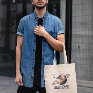 May include: Beige tote bag featuring a graphic of Pluto with an American flag design and the text "NEVER FORGET PLUTO 1930-2006." The bag is held by a person wearing a denim shirt and black shirt.