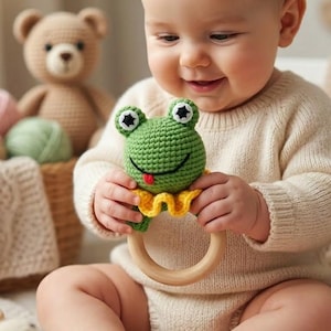 Könnte beinhalten: Ein lächelndes Baby hält eine grüne gehäkelte Froschrassel mit gelbem Kragen und Holzring. Der Frosch hat schwarz-weiße Augen und eine rote Zunge. Ein brauner Teddybär und ein Korb befinden sich im Hintergrund.