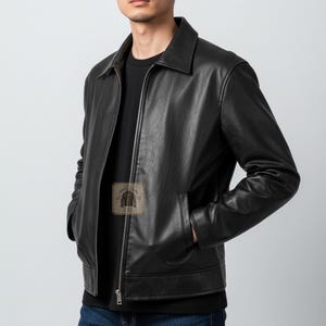 May include: A black leather jacket with a classic collar, a full-length front zipper, and two side pockets. The jacket is worn over a plain black t-shirt and blue denim jeans. A small square logo patch with the text Rogue Cuts is visible.