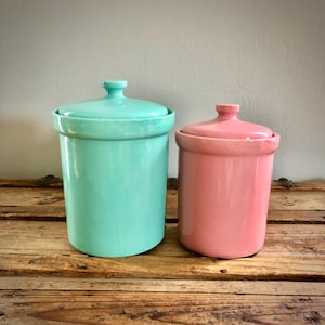 Puede incluir: Dos botes cilíndricos de cerámica con tapas a juego. El bote más grande es de color turquesa brillante, mientras que el más pequeño es de un suave rosa pastel. Ambos recipientes presentan un acabado liso y brillante con un diseño sencillo.