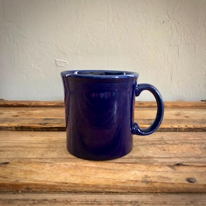 May include: A solid cobalt blue ceramic mug with a smooth, glossy finish. The mug features a cylindrical body with a slightly flared rim and a curved handle on the right side. The design is simple and functional with a deep, uniform blue color.